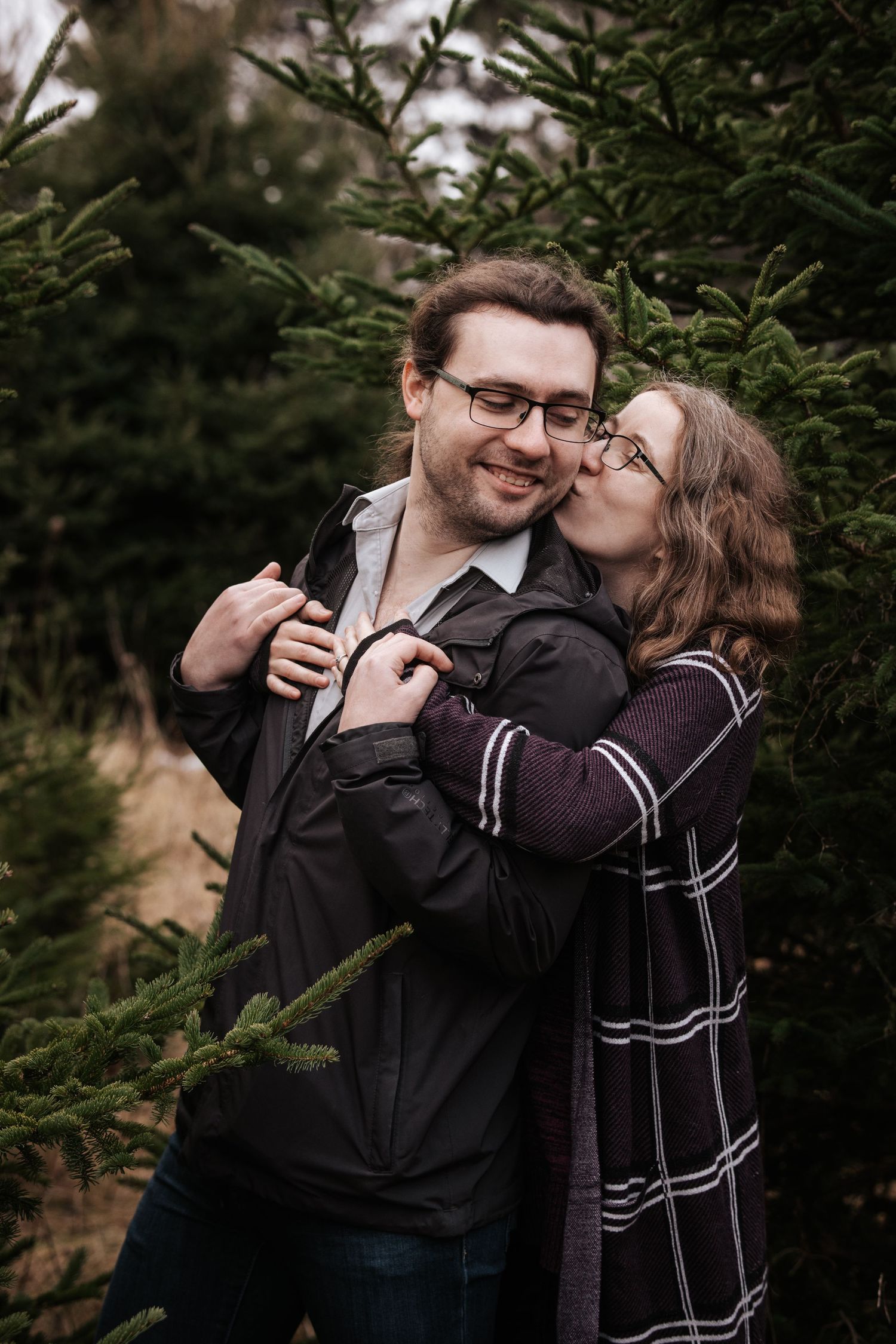 A couple shares intimate moments while standing among evergreen trees in a romantic outdoor photoshoot.