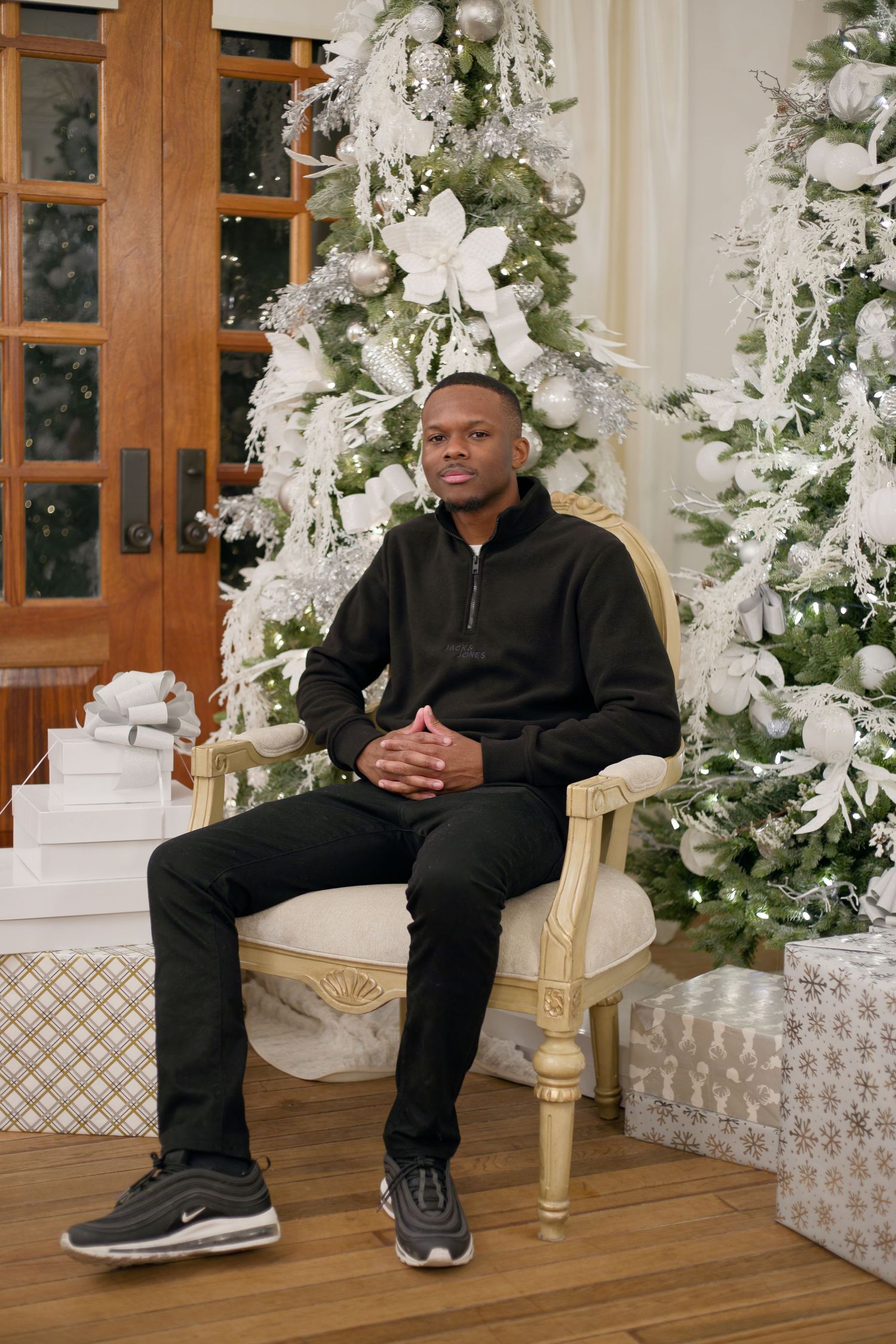A young Black man in all-black attire sits in an ornate chair beside a decorated white Christmas tree.