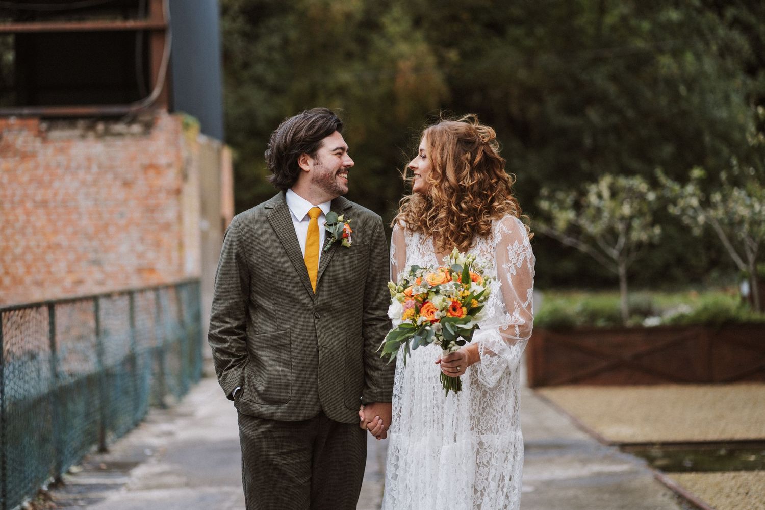 A couple in formal wedding attire poses against a rustic brick wall and wooden door backdrop.