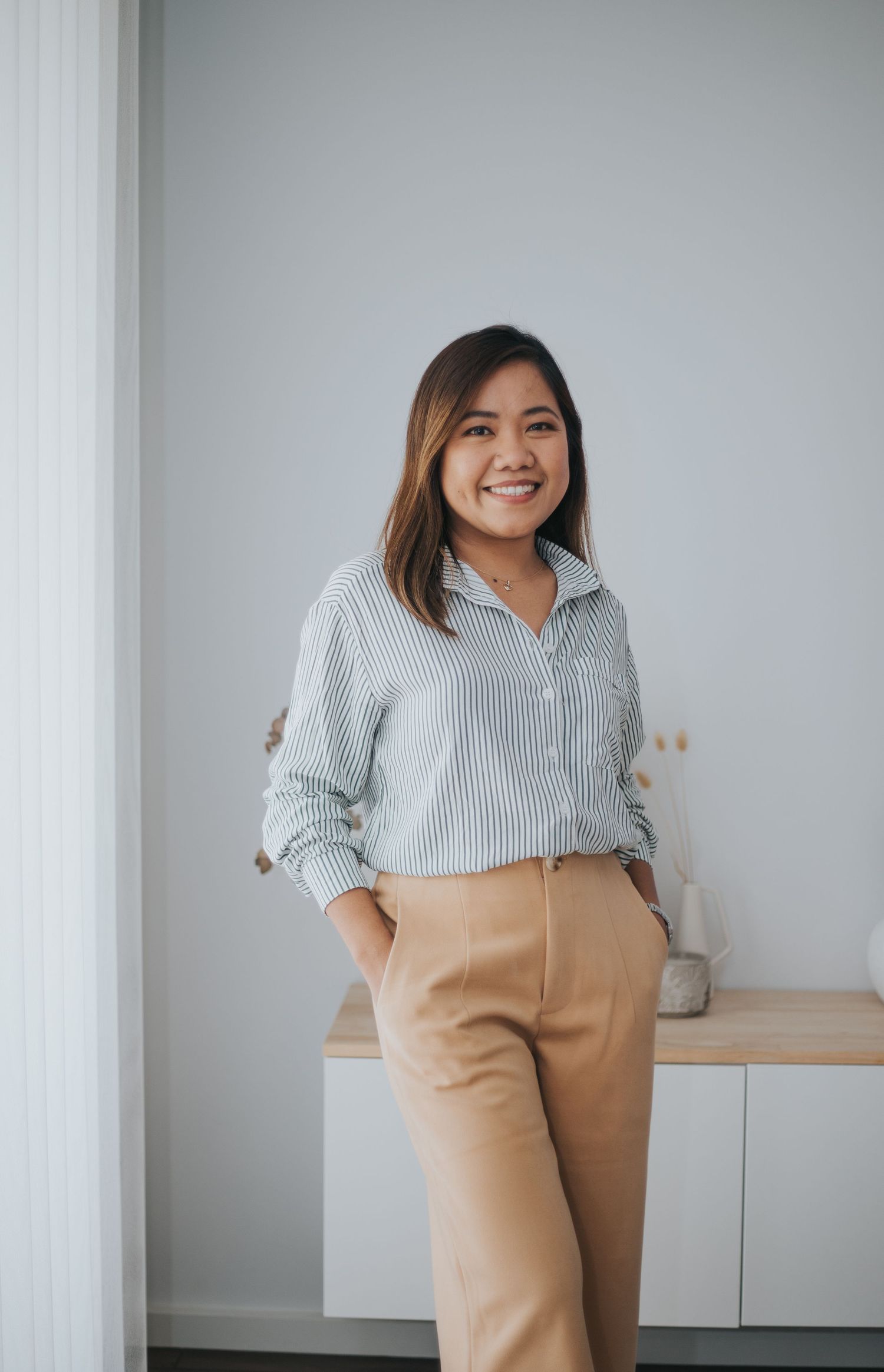 Smiling young Asian woman in striped shirt and tan trousers standing in a bright modern room.