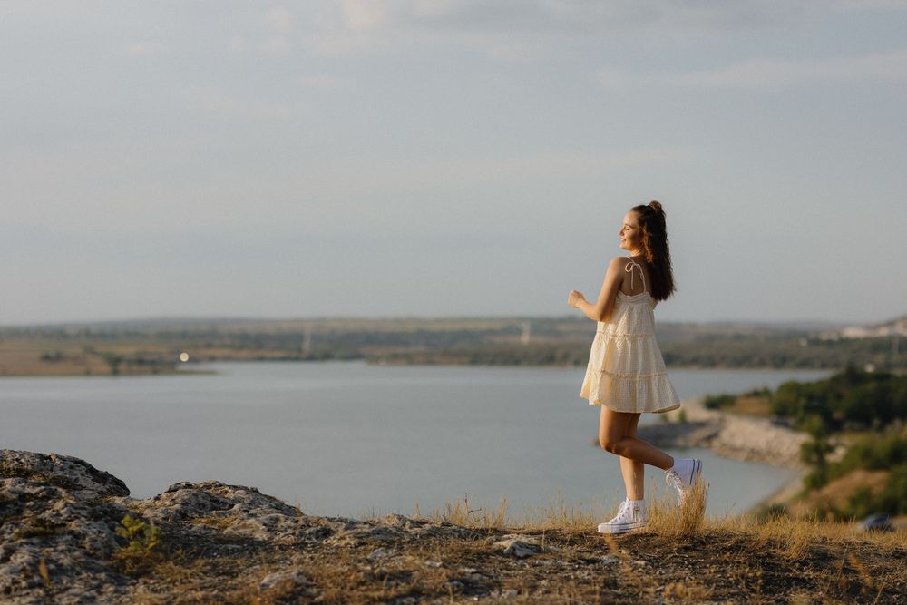 A person in a white dress stands on a rocky overlook gazing at a scenic coastal landscape during golden hour.