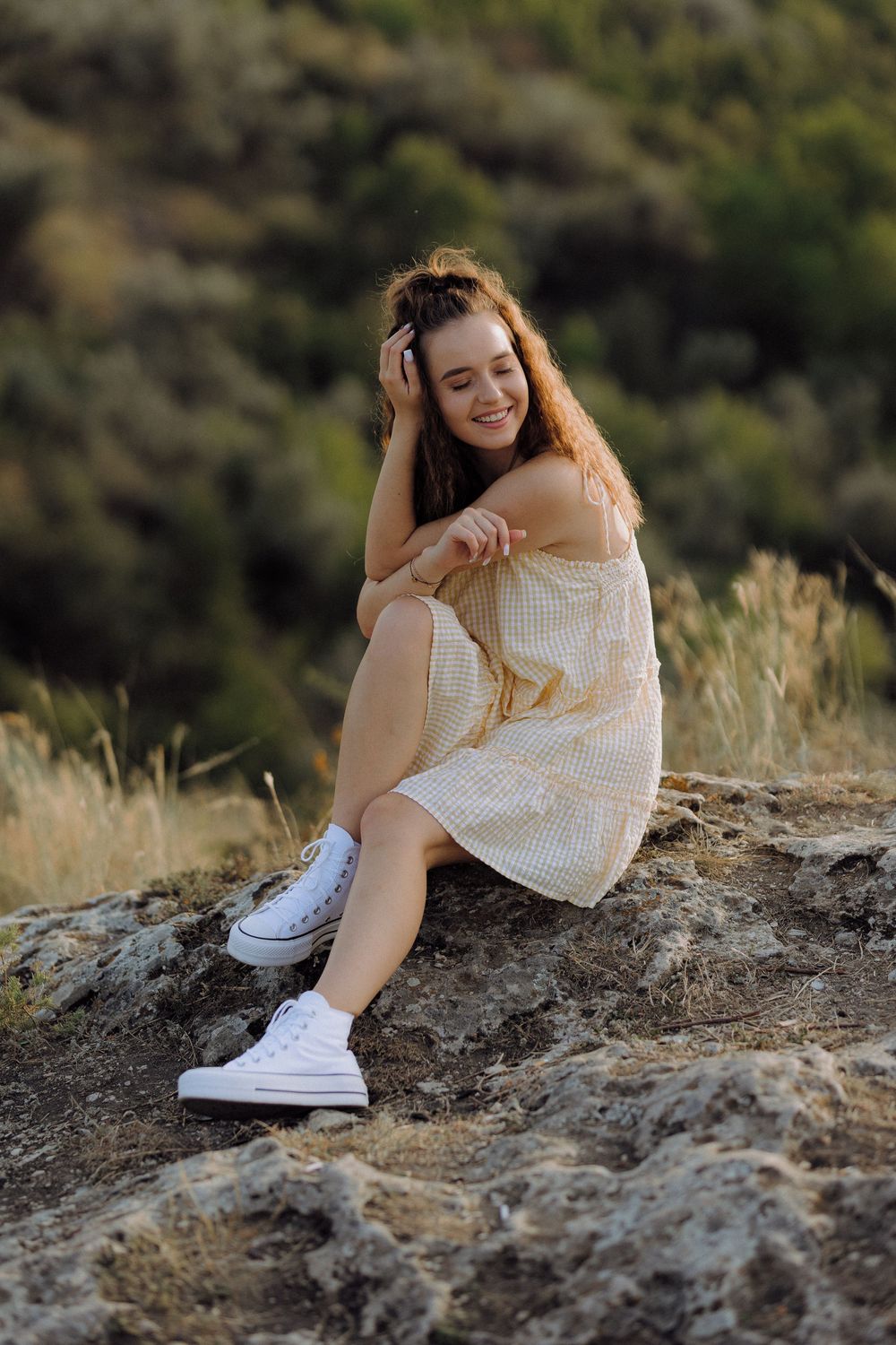 A candid outdoor portrait shows someone in a white sundress and sneakers sitting on rocky terrain at sunset.