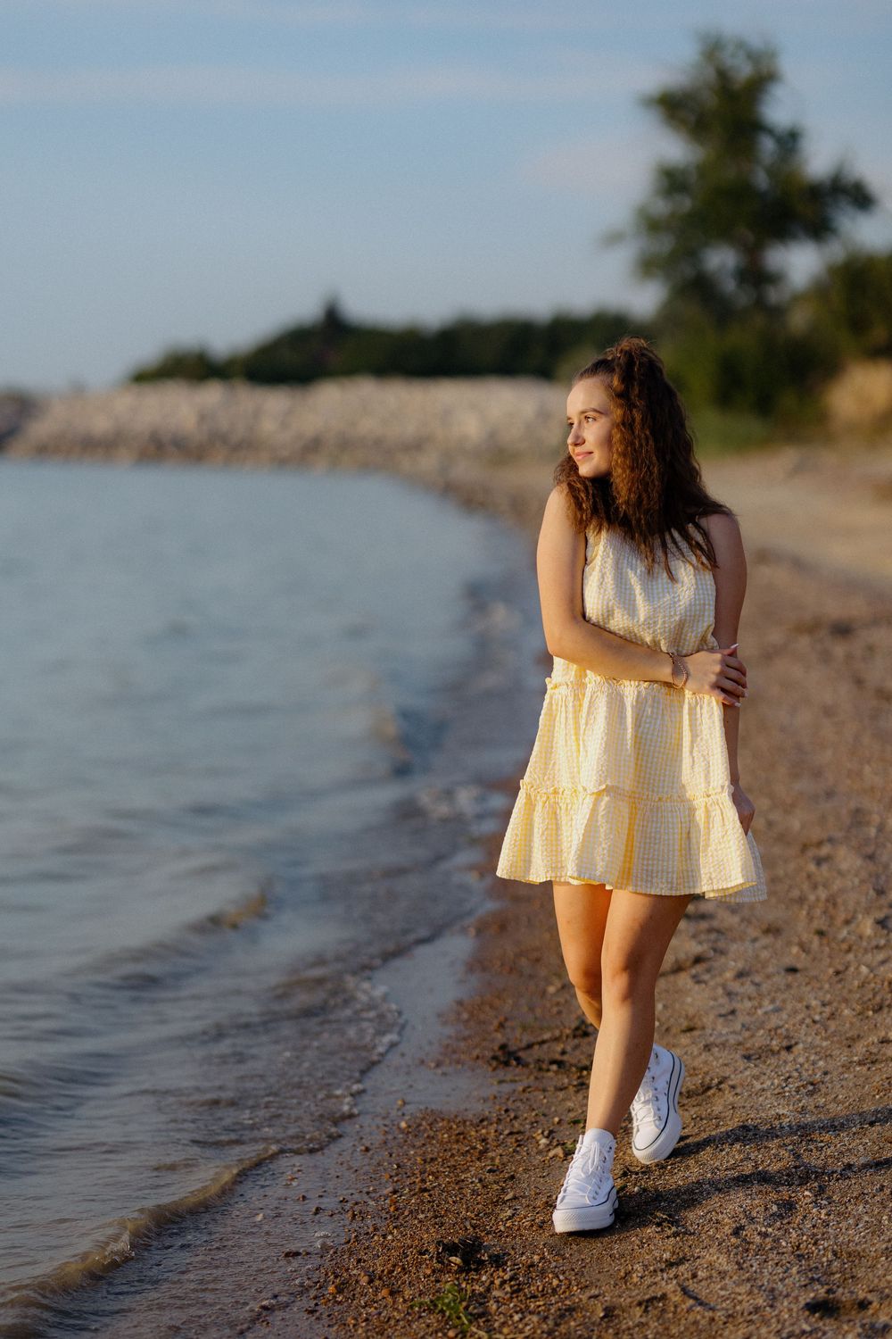 Someone in a yellow summer dress and white sneakers walks along a sandy beach at sunset.