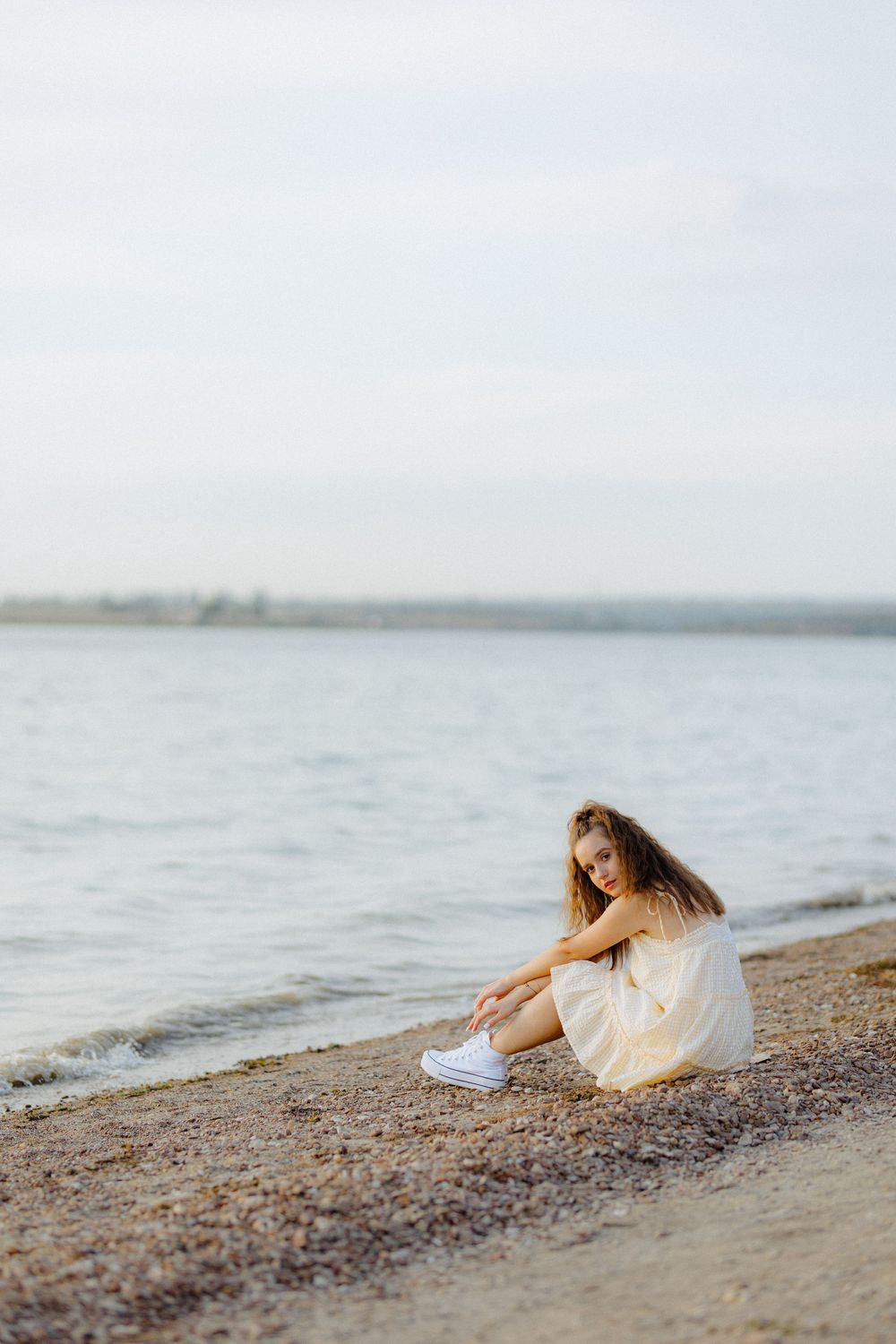 A figure in a white dress sits contemplatively on a rocky beach shore at sunset.