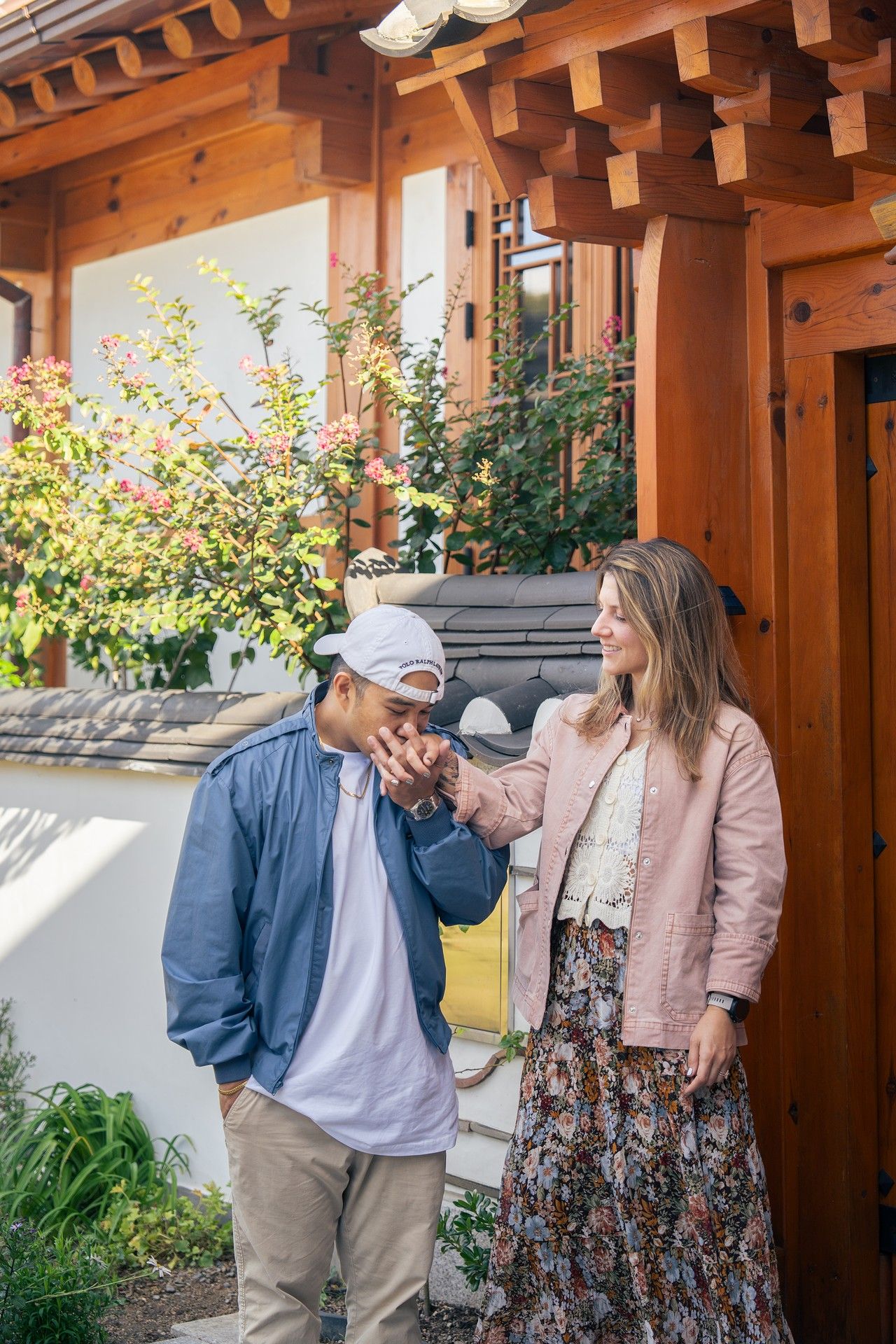 A couple in casual attire shares a playful moment under a wooden pergola surrounded by greenery.