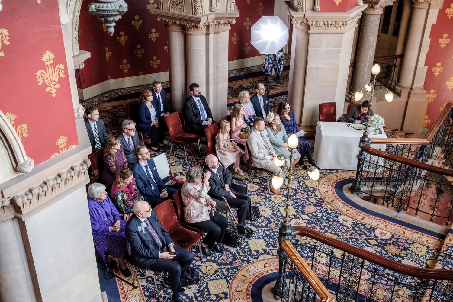 Wedding guests are seated in an elegant historic venue with ornate columns and decorative details.