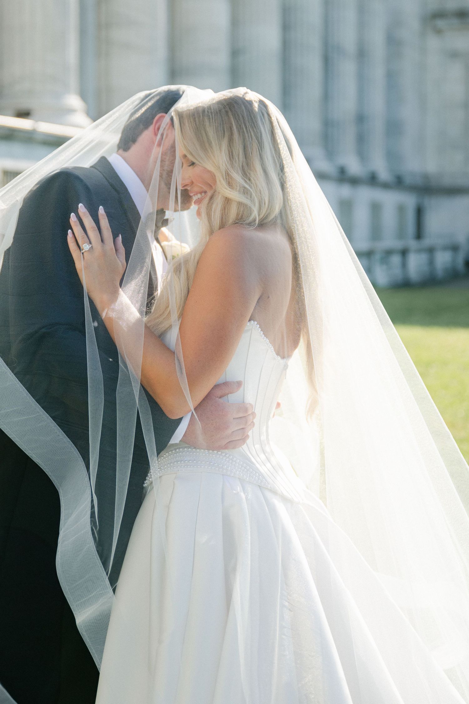 A romantic wedding couple shares their first kiss under a flowing white veil in an outdoor garden setting.