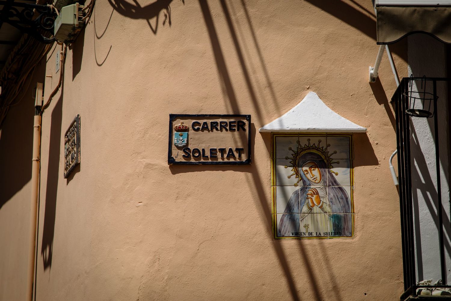 Street sign and religious icon mounted on beige wall in sunlight.