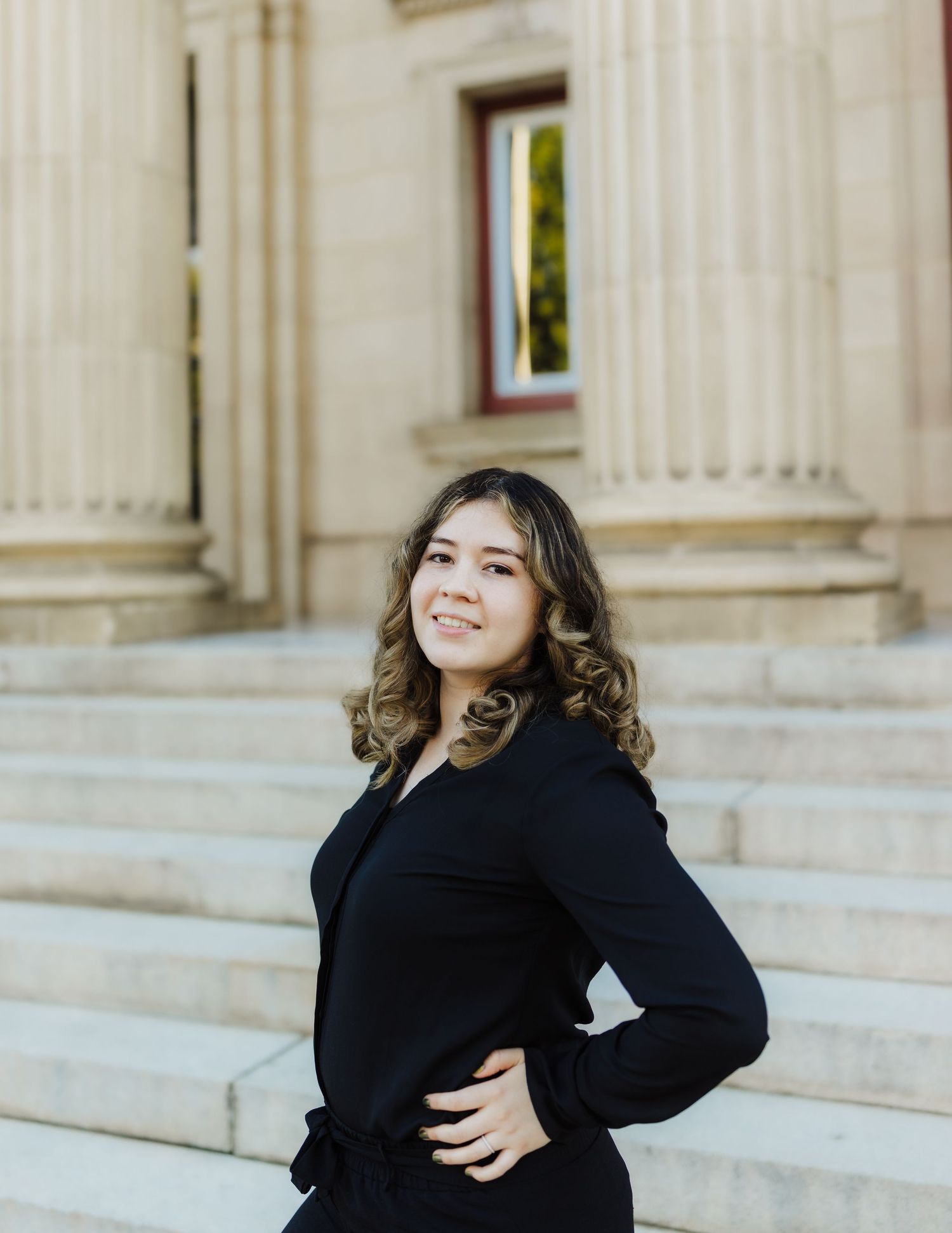 A person in a black sweater poses confidently on marble steps in front of a classic columned building.