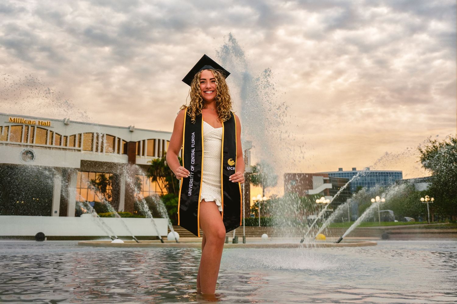 Graduate in cap and gown stands in a fountain during sunset celebration photoshoot.