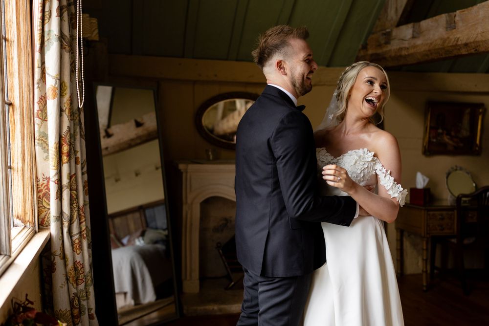 Happy couple sharing a joyful moment near a window in their wedding attire.