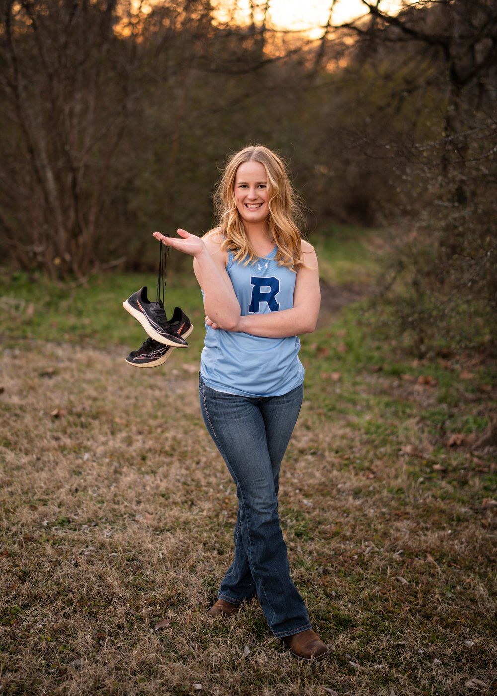 A woman in a blue tank top and jeans dances playfully in a natural outdoor setting at sunset.