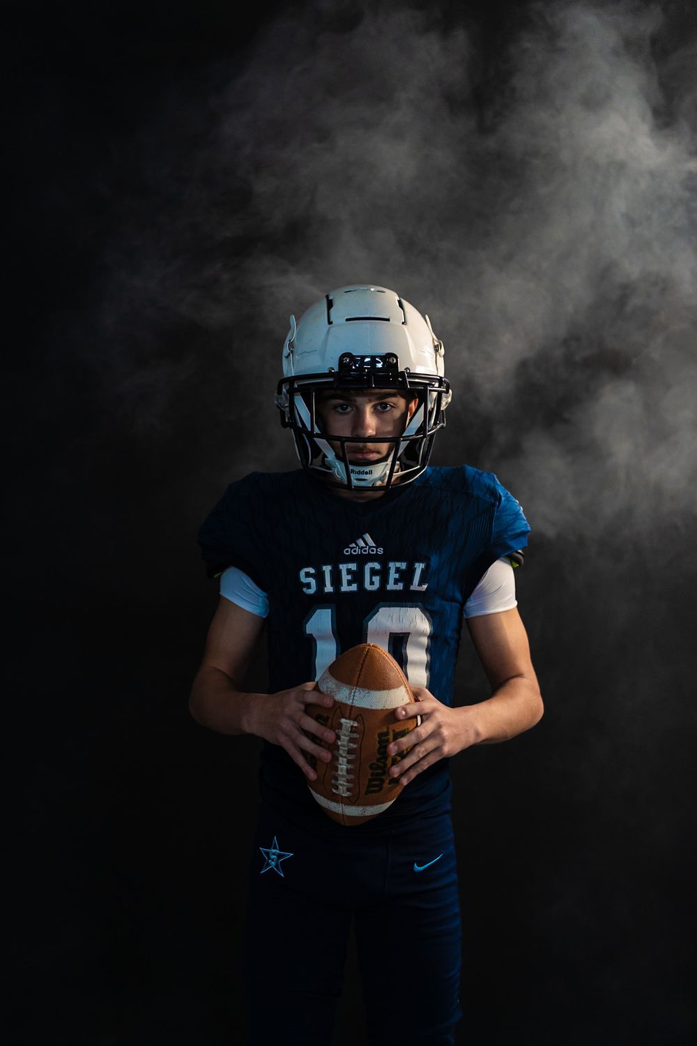 Football player in navy blue uniform holds ball against dramatic dark smoky background in dramatic sports portrait.