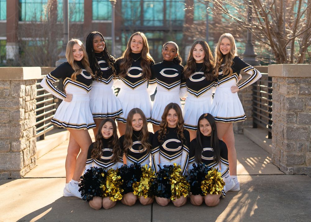 A cheerleading squad poses together outdoors in matching black uniforms with yellow pom poms.