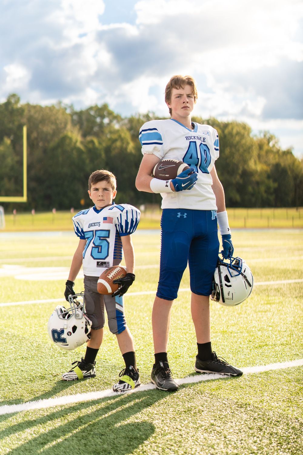 Youth football players stand in line on sunny field wearing blue and white uniforms during practice.