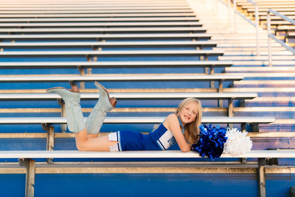 A cheerleader in a blue uniform poses on stadium bleachers with pom poms on a sunny day.