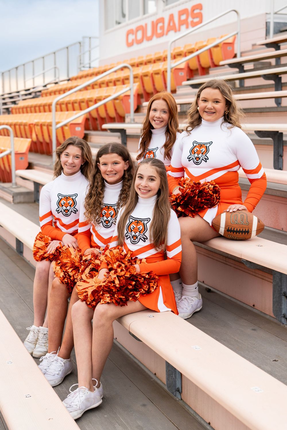 Cheerleaders in orange and white uniforms with tiger mascot logos sit on bleachers at a Cougars stadium.