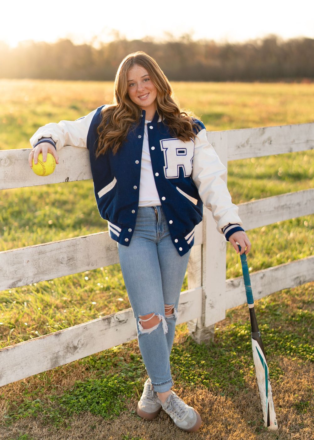 A varsity letterman jacket and ripped jeans outfit styled against a rustic white fence at golden hour sunset.