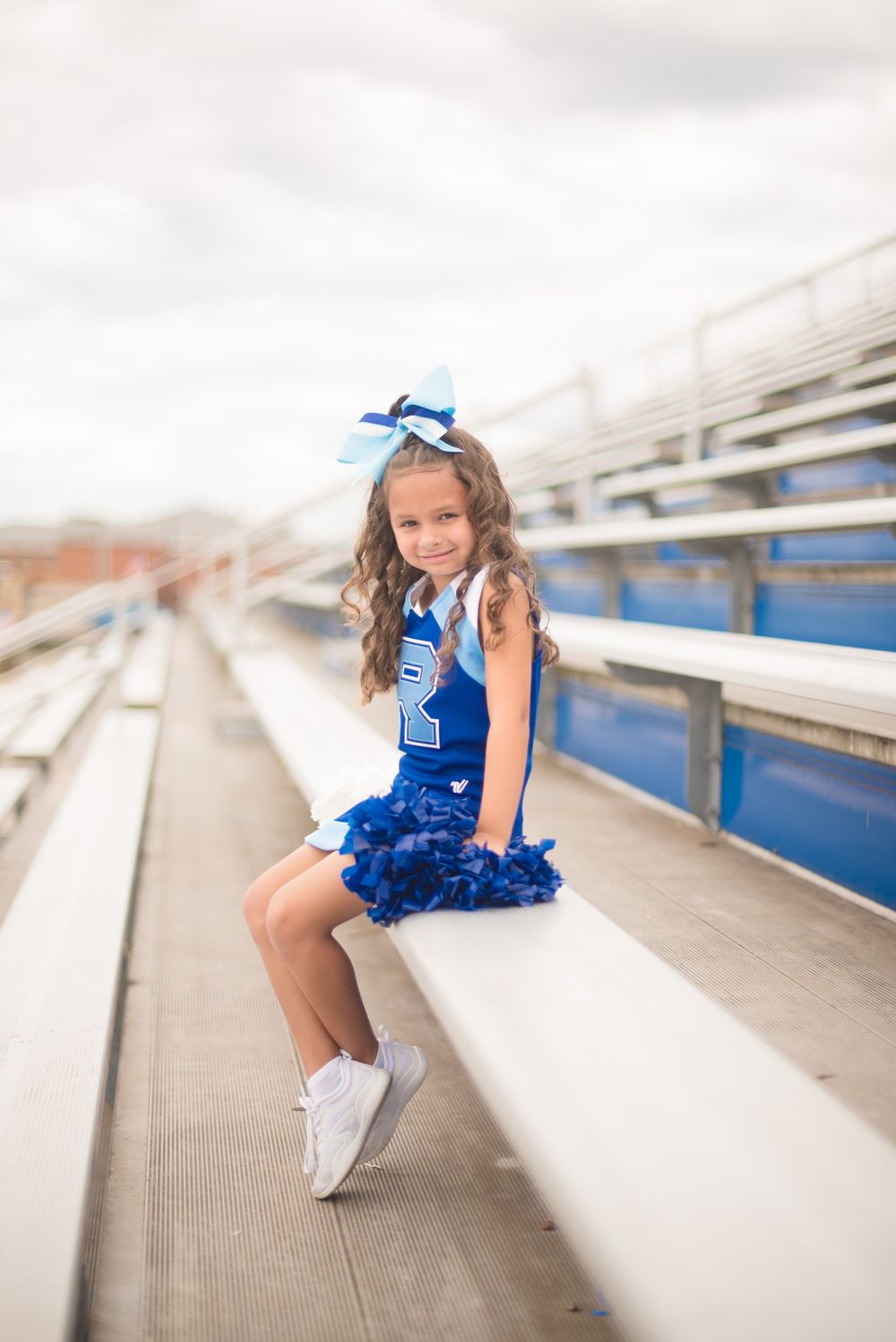 A Rockvale Youth cheerleader in a blue uniform and bow sits on stadium bleachers wearing white sneakers holding pom poms.