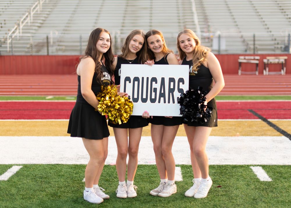 Christiana Middle School Cheerleaders in black uniforms hold Cougars sign while posing on football field with stadium seating behind them.