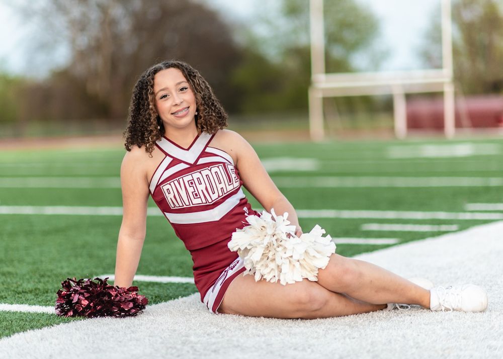 A Riverdale High School cheerleader in a burgundy red uniform poses with pom poms on a football field's sideline.