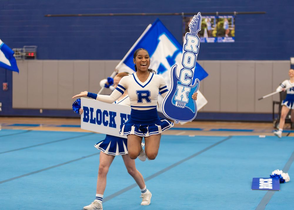 Cheerleaders in blue and white uniforms perform synchronized jumps during a routine at an indoor sports venue.