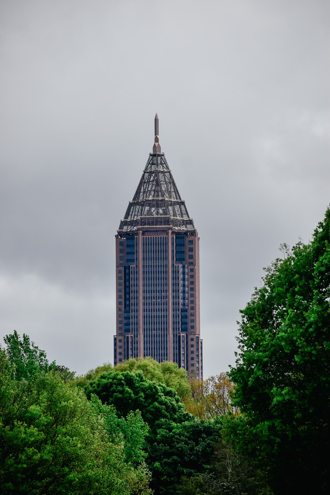 Tall modern skyscraper with distinctive pointed spire rising above trees against cloudy sky.