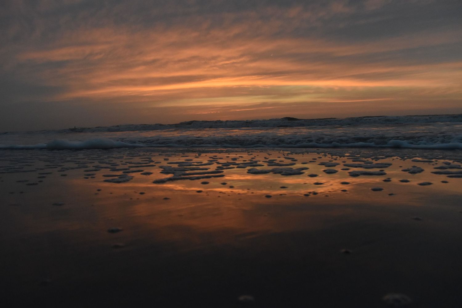 Dramatic orange sunset reflects on wet sand at a beach with waves in the distance.