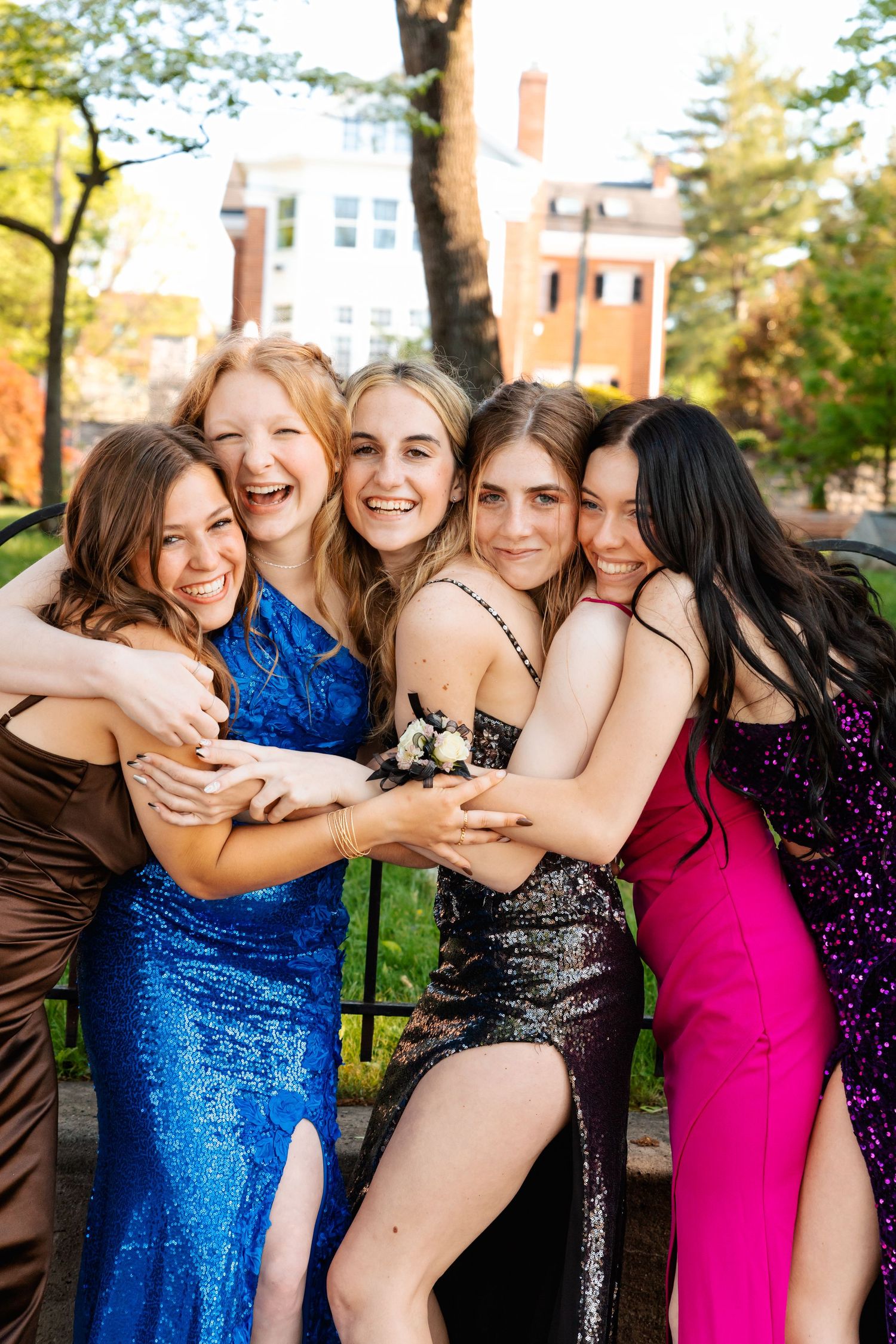 Group of friends in sparkly formal dresses hugging and laughing together at outdoor event.
