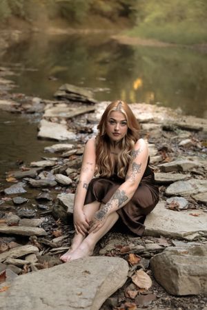 Woman in brown dress sitting on rocky riverbank with tattoos and long blonde hair.