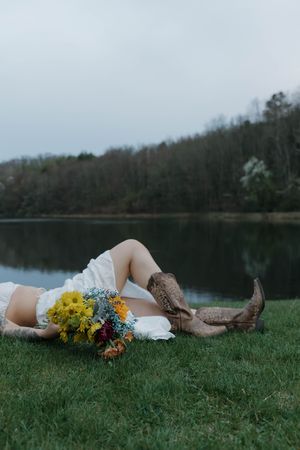Someone in cowboy boots and a white dress lies on grass by a tranquil lake holding a bright yellow wildflower bouquet.