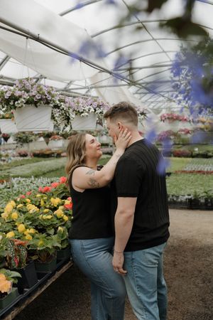 Romantic couple sharing intimate moment among colorful flowers in greenhouse with hanging floral arrangements.