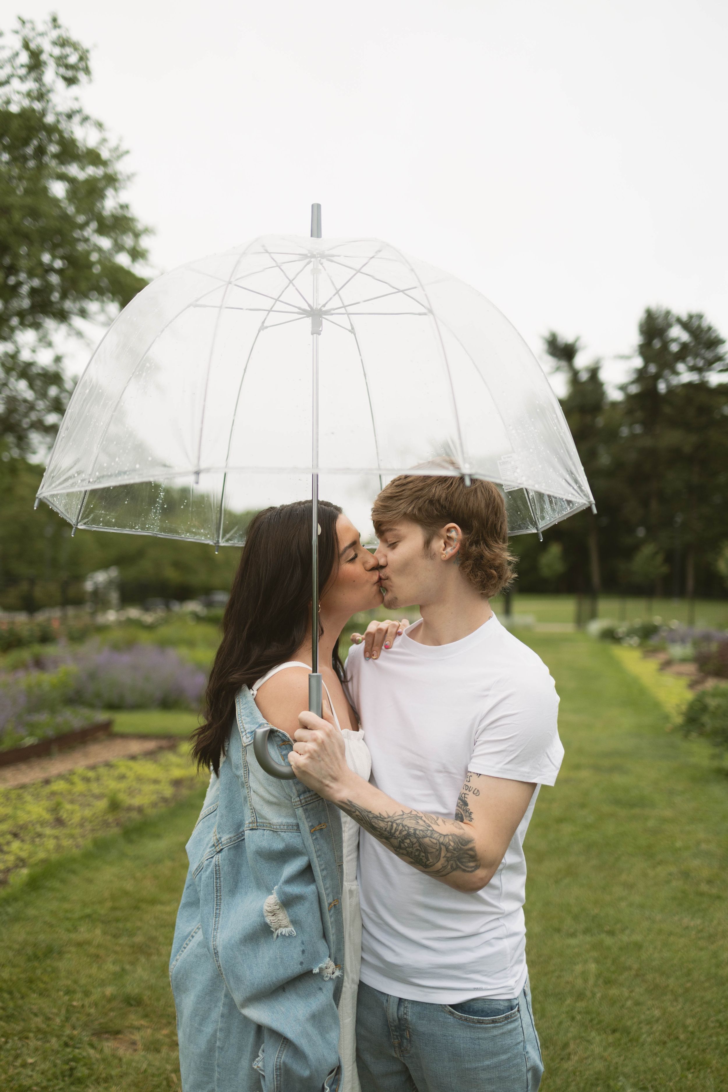 A couple shares a romantic moment under a clear umbrella in a garden setting with greenery in the background.