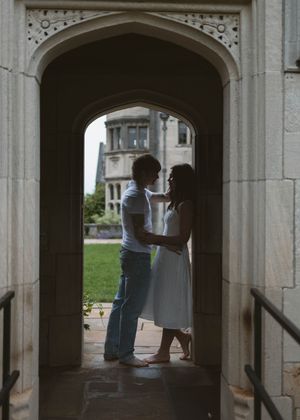 A silhouetted couple shares an intimate moment under a Gothic stone archway at dusk, with historic architecture visible beyond.