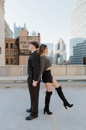 Two people stand back-to-back on a rooftop with Pittsburgh skyline in background, contrasting formal and casual outfits.