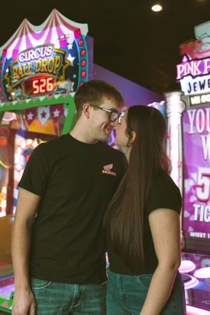 A couple wearing matching black t-shirts enjoying a romantic moment at an arcade with colorful neon lights and games.