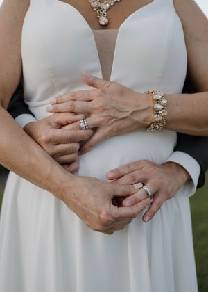 A couple wearing wedding rings and elegant jewelry embrace in a tender moment wearing white formal attire.