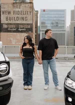 Two people sit on the edge of an urban rooftop parking garage with city buildings visible in the background.