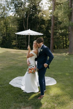 A couple in wedding attire dances playfully on a grassy hill with trees in the background during golden hour.