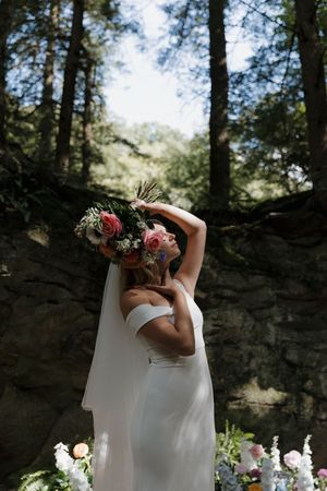 A figure in a white wedding dress holds a vibrant bouquet of red and white flowers against a rocky forest backdrop.