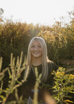 A person with long blonde hair stands outdoors during a golden sunset among tall bushes and plants in a natural setting.
