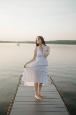 A person in a flowing white dress stands barefoot on a wooden dock over calm water at sunset.