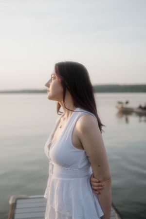 A person in a white summer dress stands on a dock at sunset, overlooking a peaceful lake with boats in the distance.