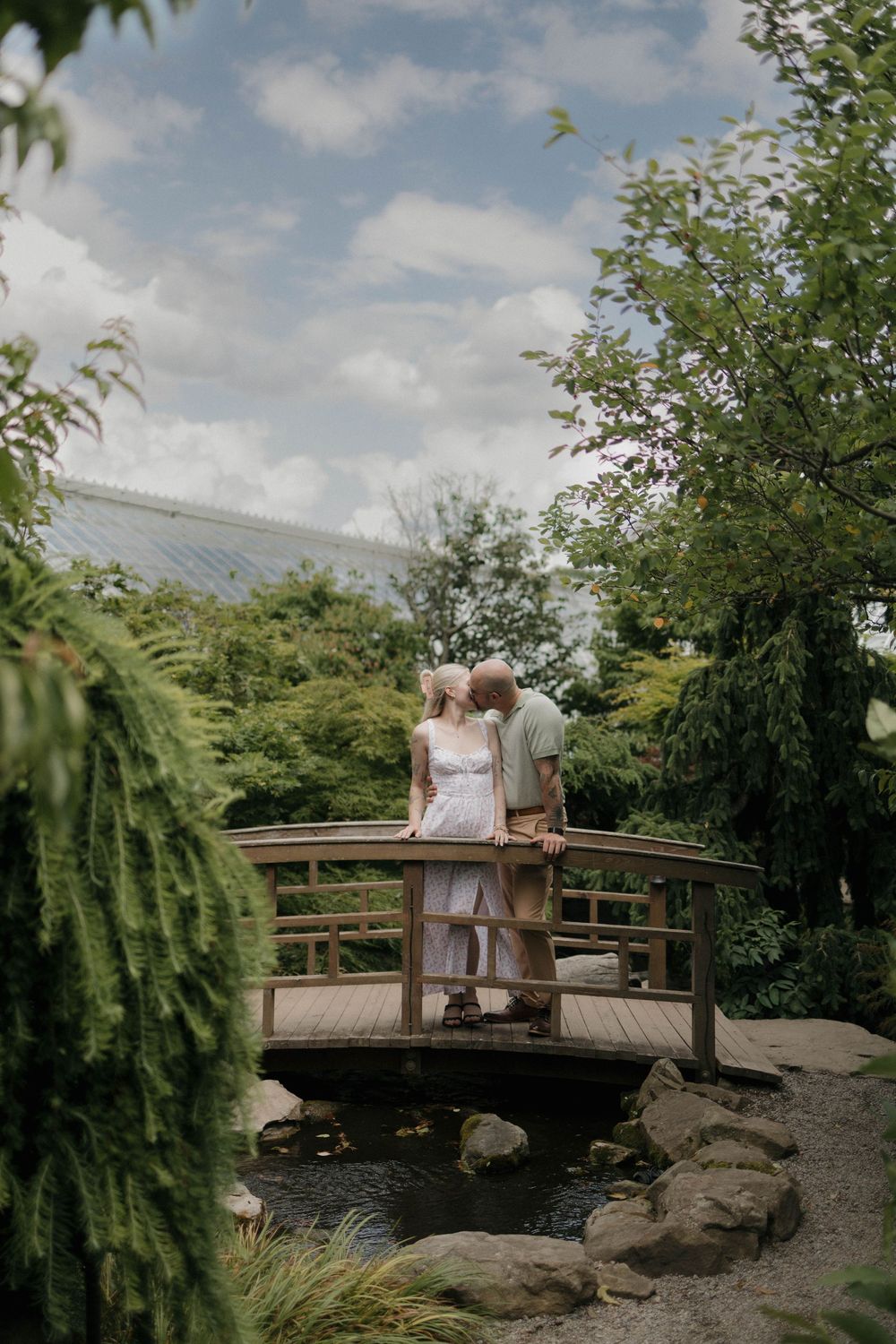 Couple standing on a wooden bridge in a lush garden, sharing a romantic moment surrounded by greenery.