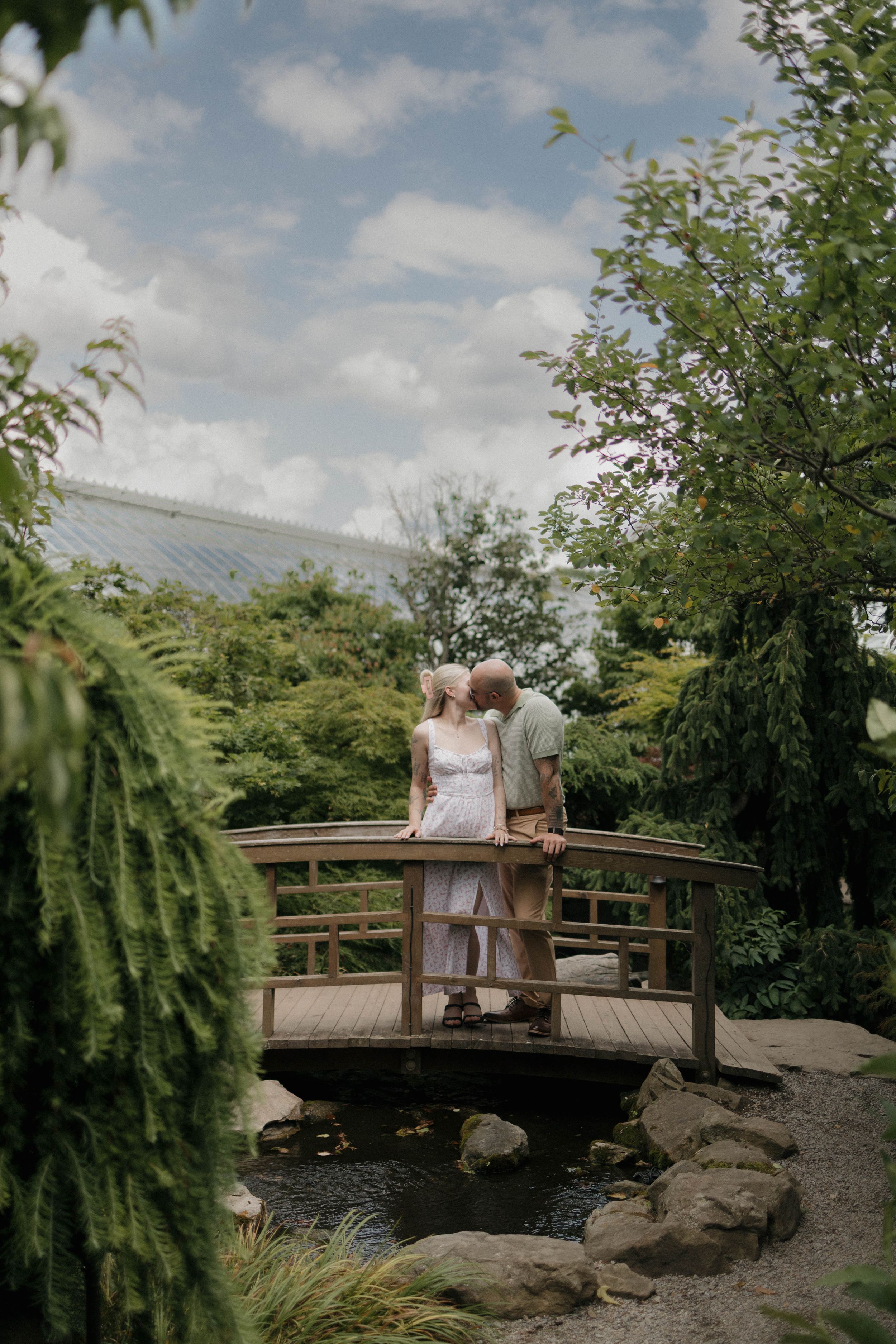 Couple standing on a wooden bridge in a lush garden, sharing a romantic moment surrounded by greenery.