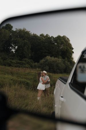 A rear view mirror reflection captures two people embracing in a grassy field at twilight beside a car.