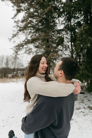 A couple embraces and laughs together while walking through a snowy winter landscape surrounded by evergreen trees.