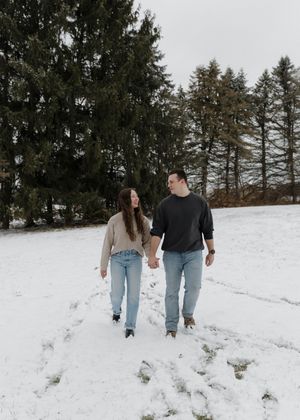 Couple walking hand in hand through a snowy winter landscape with evergreen trees in the background.