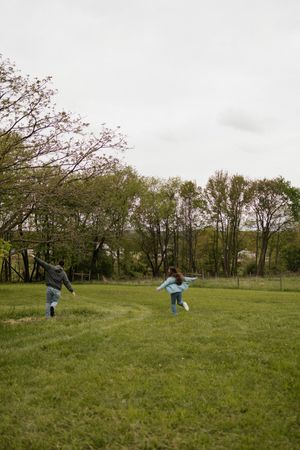 Two children running and playing in a lush green field surrounded by trees on a cloudy day.