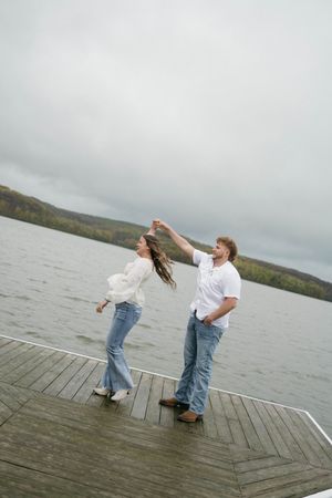 Couple dancing on a wooden dock by a lake, wearing white shirts and jeans on a cloudy day.